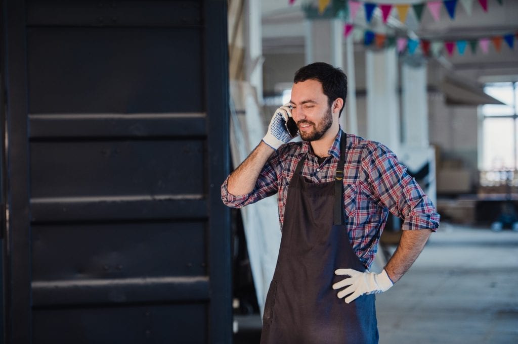 Smiling bearded man wearing gloves, owner of a small woodwork business, standing in his workshop